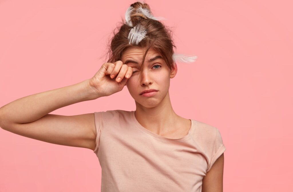 A person with feathers in their hair rubbing their dry and tired eyes.